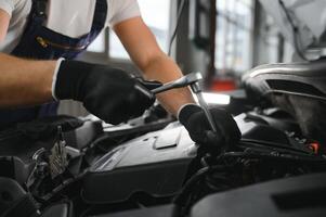 Beau Jeune Masculin auto mécanicien dans spécial uniforme vêtements en portant une lampe de poche, à la recherche pour panne et réparer en dessous de le capuche dans le voiture moteur dans une voiture atelier photo