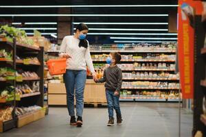 Jeune femme et sa peu fils portant protecteur visage masque magasin une nourriture à une supermarché pendant le coronavirus épidémie ou grippe épidémie. photo