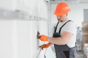 homme cloison sèche ouvrier ou plâtrier en mettant engrener ruban pour placoplâtre sur une mur en utilisant une spatule et plâtre. portant blanc casque, travail gants et sécurité lunettes. image avec copie espace. photo