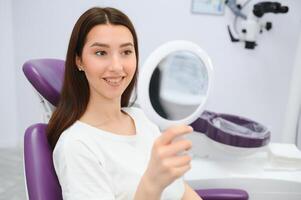 jolie fille dans dentiste cabinet est en utilisant une miroir à Regardez par le idéal travail de une médecin. photo