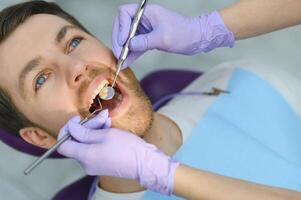 parodontal prestations de service. fermer coup de souriant homme avoir traitement dans stomatologique clinique, dentiste médecin dans gants en utilisant stérile dentaire outils pour examiner les dents de Masculin patient photo