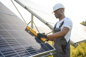 Afro-américain ouvrier dans gris salopette et une blanc difficile chapeau travaux dans une champ de solaire panneaux. solaire renouvelable énergie concept. photo