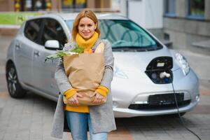 mise en charge électro voiture à le électrique gaz gare. femme permanent par le auto. Dame avec produit alimentaire photo