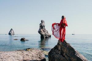 femme Voyage mer. Jeune content femme dans une longue rouge robe posant sur une plage près le mer sur Contexte de volcanique rochers, comme dans Islande, partage Voyage aventure périple photo