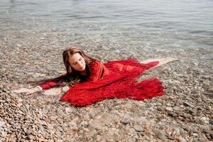 femme Voyage mer. content touristique dans rouge robe prendre plaisir prise image en plein air pour souvenirs. femme voyageur posant dans mer plage, entouré par volcanique montagnes, partage Voyage aventure périple photo