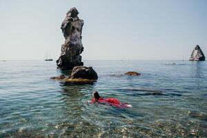 femme Voyage mer. content touristique dans rouge robe prendre plaisir prise image en plein air pour souvenirs. femme voyageur posant dans mer plage, entouré par volcanique montagnes, partage Voyage aventure périple photo