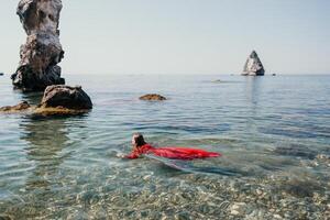 femme Voyage mer. content touristique dans rouge robe prendre plaisir prise image en plein air pour souvenirs. femme voyageur posant dans mer plage, entouré par volcanique montagnes, partage Voyage aventure périple photo