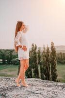 content femme dans blanc boho robe sur le coucher du soleil dans montagnes. romantique femme avec longue cheveux permanent avec sa retour sur le le coucher du soleil dans la nature dans été avec ouvert mains. silhouette. la nature. le coucher du soleil. photo