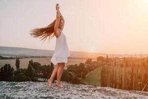 content femme permanent avec sa retour sur le le coucher du soleil dans la nature dans été avec ouvert mains. romantique magnifique la mariée dans blanc boho robe posant avec montagnes sur le coucher du soleil photo