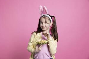 Jeune joyeux enfant posant avec Pâques des œufs et jouets sur caméra, sentiment content et excité à propos printemps vacances fête et cadeaux. peu fille avec lapin oreilles souriant dans studio. photo