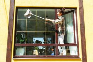 une femme manuellement lavages le fenêtre de le maison avec une chiffon avec une vaporisateur nettoyeur et une balai dehors. sécurité à hauteur, restauration commande et propreté dans le printemps, nettoyage un service photo