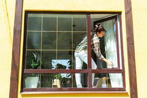 une femme manuellement lavages le fenêtre de le maison avec une chiffon avec une vaporisateur nettoyeur et une balai dehors. sécurité à hauteur, restauration commande et propreté dans le printemps, nettoyage un service photo