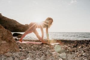 femme mer yoga. content femme dans blanc maillot de bain et boho style bracelets pratiquant en plein air sur yoga tapis par mer sur le coucher du soleil. femmes yoga aptitude routine. en bonne santé mode de vie, harmonie et méditation photo