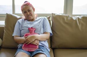 Jeune asiatique femme les usages une chaud l'eau sac à soulager menstruel crampes. elle est assis sur le canapé. photo