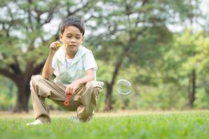 garçon séance dans le parc avec soufflant air bulle, entouré par verdure et la nature photo