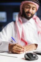 arabe homme assise à le sien Bureau table absorbé dans le sien emploi. il est travail sur une mini-ordinateur, prise détaillé Remarques et conduite en ligne recherche, mettant en valeur une dévoué et concentré approche. photo