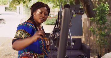 Jeune amateur de sport femelle attentivement examine bicyclette pédales et freins assurer lisse et efficace Extérieur vélo. africain américain femme cycliste Faire annuel vélo entretien. ordinateur de poche tir. photo