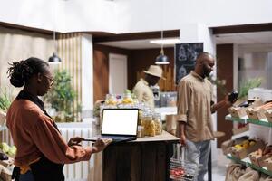 africain américain épicerie magasin propriétaire regards à portable avec une Vide blanc filtrer. Jeune femme examiner des biens tandis que en fonctionnement une sans fil PC avec chromakey fond modèle. durable, local des produits. photo