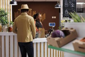 une amical femelle la caissière avec une gros sourire sur sa visage et une beau Masculin client portant une chapeau. elles ou ils sembler à être ayant une agréable interaction à le check-out compteur dans une épicerie magasin. photo