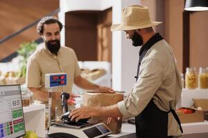 milieu est vendeur étant de un service à caucasien client à check-out comptoir. à la caissière bureau, Masculin vendeur portant une chapeau tandis que en utilisant numérique échelle à mesure articles pour le Jeune client. photo