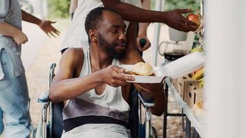 gratuit repas et des fruits sont donné à une en fauteuil roulant africain américain homme dans besoin. handicapé, pauvres et sans abri gens recevoir des provisions de une non lucratif nourriture conduire. fermer, trépied tir. photo