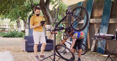 caucasien homme permanent avec intelligent dispositif tandis que africain américain femme démantèle endommagé bicyclette roue. couple en utilisant numérique tablette pour recherche de vélo ajustements pour été entretien. photo