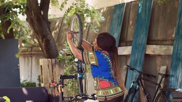 actif africain américain femme avec prudence relâchement et enlever endommagé vélo roue. amateur de sport femelle cycliste soigneusement démantèlement pneu de bicyclette de face fourchette pour entretien dans Accueil cour. photo