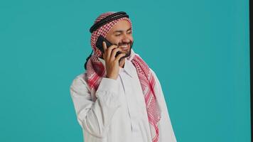 musulman homme dans traditionnel tenue envoyer des SMS sur mobile téléphone tandis que posant fièrement dans studio. milieu est adulte habillé dans thobe et à carreaux foulard, Envoi en cours en ligne messages sur téléphone. photo