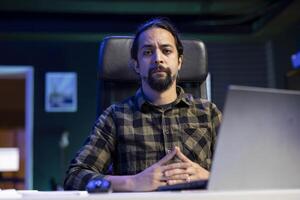 portrait de une gentilhomme assise à une bureau avec le sien mains franchi et à la recherche à caméra. sérieux Masculin individuel travail de maison, vérification e-mails, recherche sur le sien portable. photo