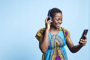charmant content femme dansant et écoute à sensationnel la musique sur écouteurs, en utilisant téléphone intelligent à changement Chansons de une playlist. africain américain fille ayant amusement avec moderne airs dans studio. photo