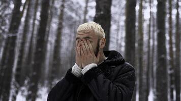 élégant branché homme dans pin arbre hiver forêt frottement le sien du froid rouge visage avec le sien mains. médias. portrait de une homme en plein air dépenses temps seul dans hiver les bois. photo