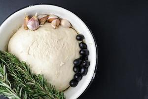 pâte crue dans un bol blanc, brins de romarin, olives et ail sur fond sombre. photo