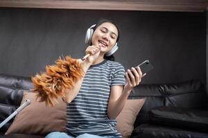 Jeune femme asiatique prendre plaisir nettoyage Ecoutez la musique écouteurs chanter plumeau brillant maison photo