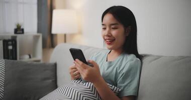 content Jeune femme séance sur canapé tenir une café agresser et utilisation mobile téléphone pour en ligne achats sans argent dans vivant pièce à Accueil ,technologie argent portefeuille et en ligne Paiement concept photo