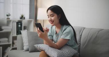 content Jeune femme séance sur canapé prendre plaisir en utilisant mobile téléphone pour en ligne achats sans argent dans vivant pièce à maison, La technologie argent portefeuille et en ligne Paiement concept photo