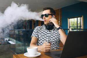 Jeune Beau branché homme lunettes de soleil séance dans café avec une tasse de café, vapoter et communiqués une nuage de vapeur. travail à ordinateur portable, parlant mobile téléphone et ayant une peu Pause. avec copie espace. photo