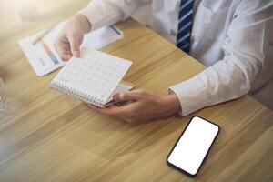 professionnel dans une blanc chemise Planification dans une journal intime avec une téléphone intelligent avec écran maquette sur le bureau pour rapide accès photo