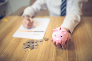 la personne dans une affaires chemise économie argent dans une porcin banque, avec pièces de monnaie et financier rapports sur le tableau. économie argent affaires concept photo