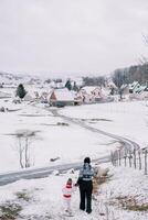 mère et une peu fille marcher vers le bas une neigeux colline en portant mains vers une village route. retour vue photo