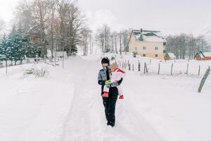 mère avec une peu fille dans sa bras des promenades le long de une neigeux village route en dessous de chute de neige photo