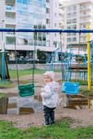 peu fille des stands près le balançoire dans de face de une flaque, tournant autour. retour vue photo