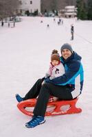 souriant papa avec une peu fille dans le sien bras est assis sur une traîneau sur une neigeux plaine photo