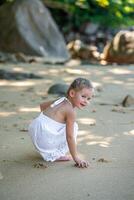 peu fille dans blanc robe ayant amusement sur rivage dans le ombre de des arbres et palmiers. haute qualité photo