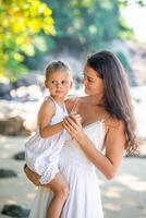 Jeune femme mère avec une peu fille dans blanc Robes sur rivage dans le ombre de des arbres et palmiers. haute qualité photo