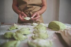 détails sur le mains de une femme femme au foyer, chef dans beige tablier fabrication Dumplings de vert pâte avec fondu épinard, et remplissage il avec en purée patates, permanent à cuisine table dans rural maison photo