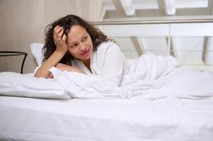 Jeune content femme mensonge dans blanc lit sur blanc oreiller et couvert avec confortable doux lit feuilles, souriant à la recherche de côté photo