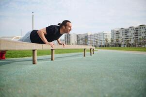 Jeune athlétique homme dans tenue de sport, Faire bar des pompes sur barre transversale, exercice en plein air dans le Urbain terrain de sport. photo