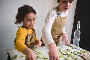 garçon et fille fabrication Dumplings, permanent à fariné cuisine table dans le rustique cuisine intérieur. cuisine classe pour enfants. en train de préparer Ravioli, pelmenis, varennyky selon à traditionnel recette photo