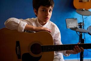 Beau adolescent garçon en jouant guitare dans le moderne la musique studio, à la recherche en toute confiance à caméra, séance contre une bleu mur avec électrique tambours. personnes. musique. loisirs et loisir photo