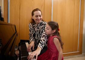 de bonne humeur mère et fille pianistes en jouant piano ensemble, performant une mélodie pour Noël pendant la musique leçon ensemble. mignonne bébé fille dans élégant rouge robe, écoute à sa maman en jouant pianoforte photo